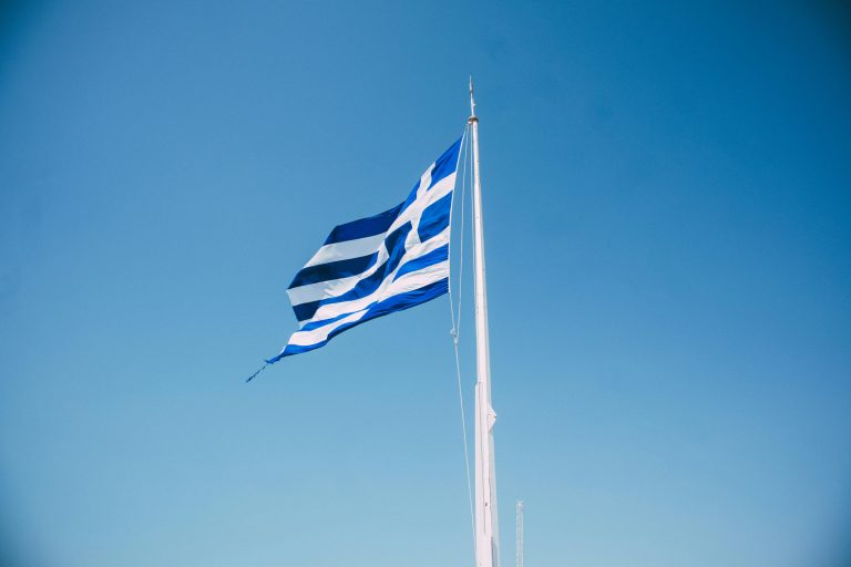The national flag of Greece waving prominently against a bright blue sky, symbolizing national pride.