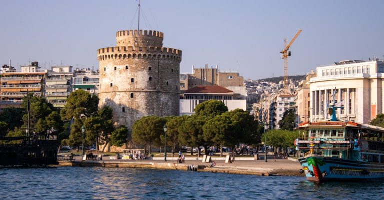 View of the iconic White Tower in Thessaloniki, Greece, by the waterfront with boats and city skyline.